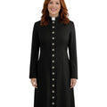 A smiling woman with red hair wearing a long black cassock with a white collar and a vertical row of silver buttons down the front, photographed against a plain white background.