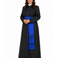 A woman modeling a long black cassock with a bright royal blue sash, blue decorative buttons, and a white collar, photographed against a plain white studio background.