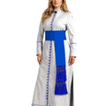 A woman modeling a long white or silver-gray traditional cassock with a bold royal blue sash and matching blue fringe, photographed against a plain white studio background.
