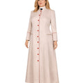 A woman modeling a long, light beige or cream-colored cassock with a high stand collar, red button front, and red piping details on the cuffs and front opening, paired with silver heels against a plain white background.