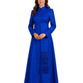 A woman modeling a long-sleeved royal blue cassock with a mandarin collar, button front, and decorative tassel details on the skirt, set against a plain white background.
