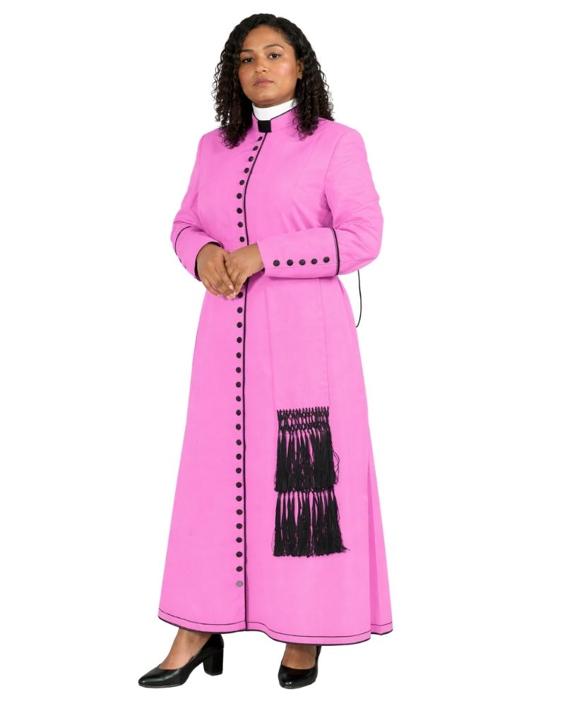 A woman modeling a bright pink cassock with black fringe and black decorative buttons, styled with a white collar and black heels, against a plain white background.