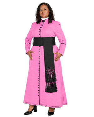 A woman modeling a bright pink long-sleeve cassock with a wide black waist sash, black fringe detail, and black decorative buttons, paired with a white collar and black shoes, against a plain white background.
