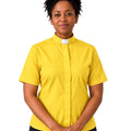 A woman wearing a bright yellow short-sleeve clerical shirt with a white collar tab, paired with black trousers, standing against a plain white background.