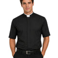 A man wearing a black short-sleeve clerical shirt with a white collar tab, paired with black trousers and a black belt, against a plain white background.